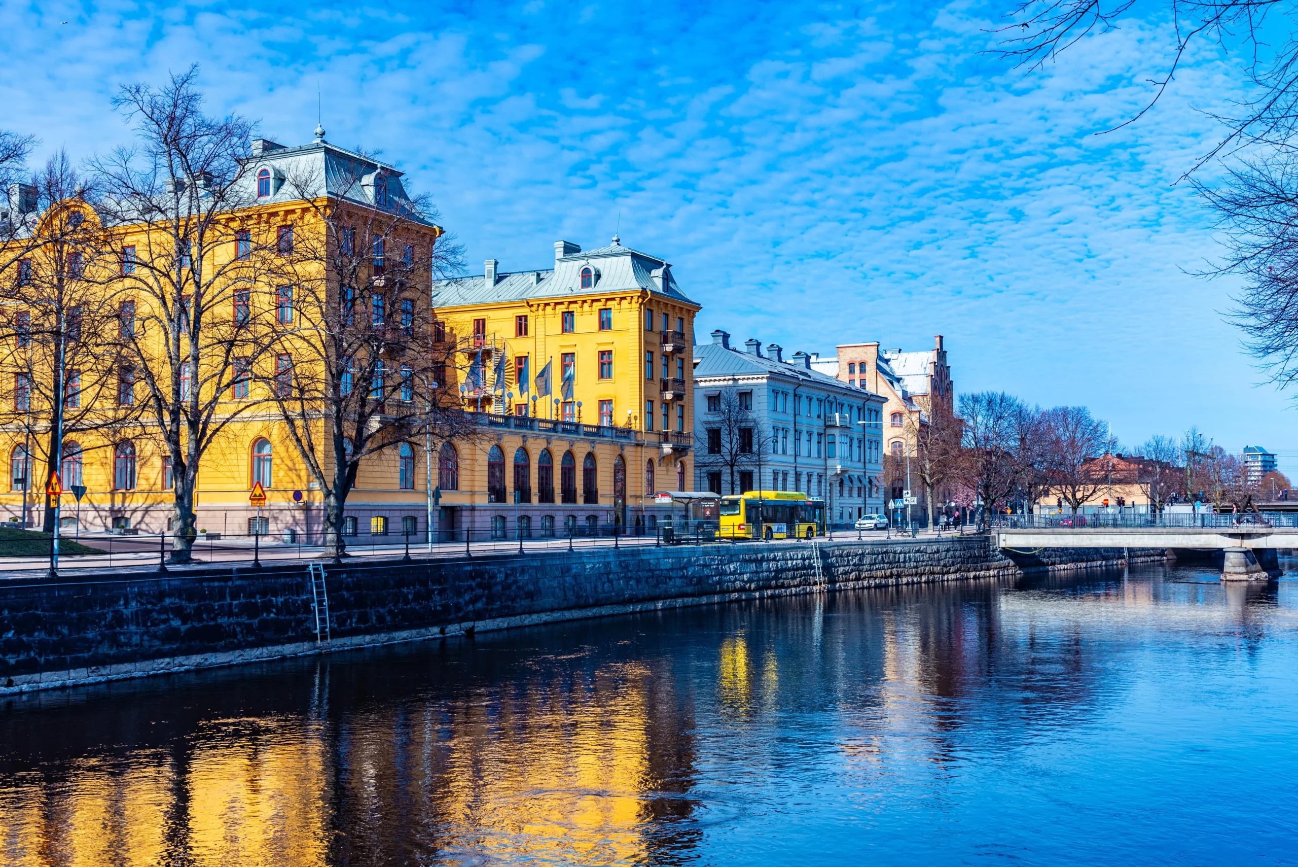 Stadsvy över centrala Västerås med gula och blå byggnader, buss, träd och promenadstråk längs vattnet.