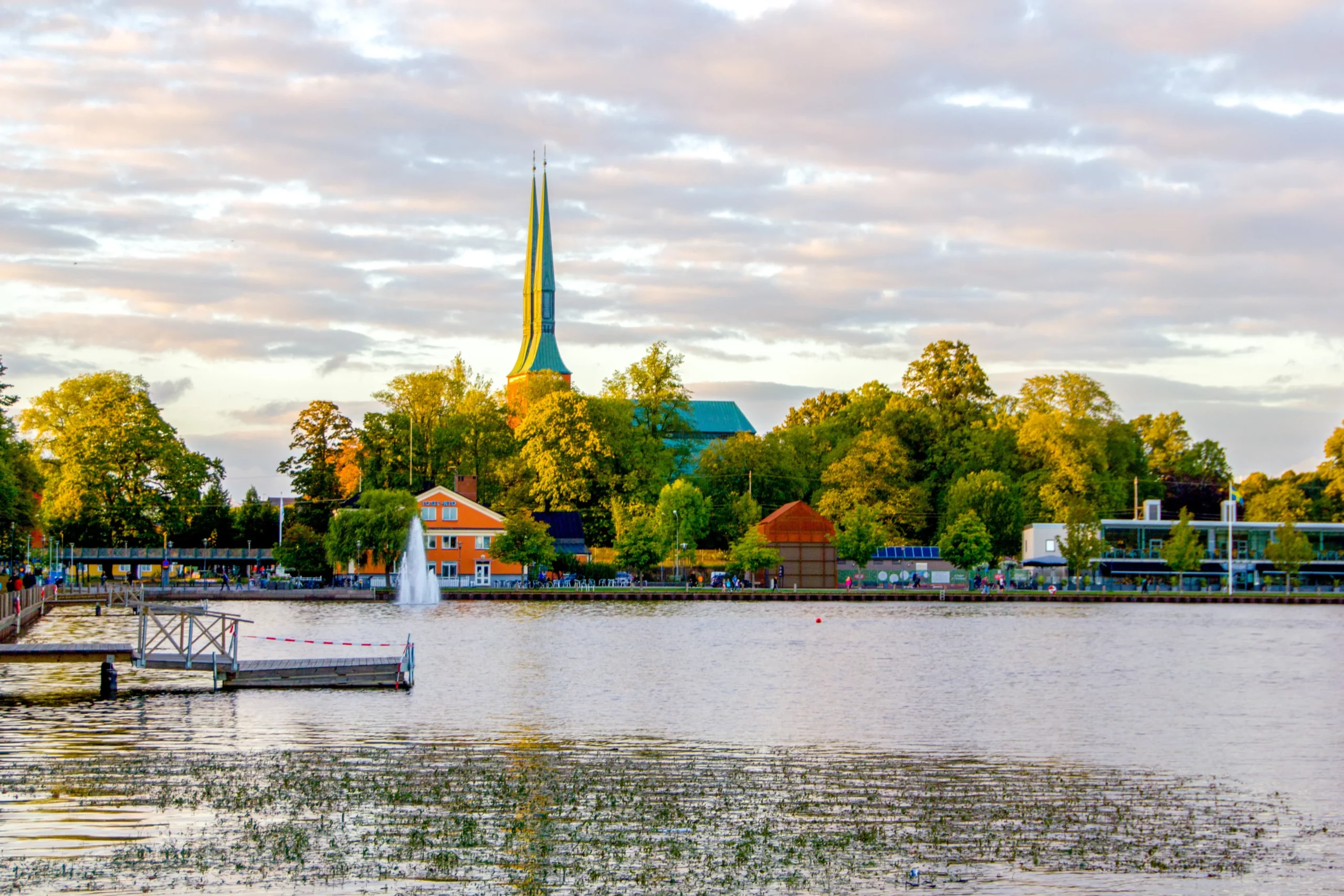 Växjö sjöpark med fontän, promenadstråk och lummiga träd i kvällssol.